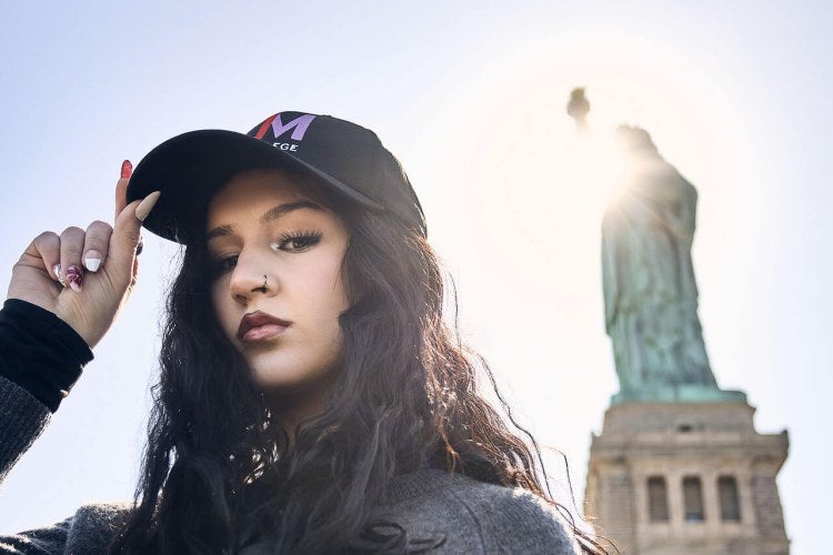 young woman, dark hair, LIM cap, stands in front of the Statue of Liberty in New York Harbor
