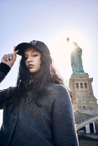 young woman, dark hair, LIM cap, stands in front of the Statue of Liberty in New York Harbor
