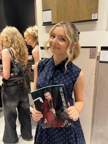 young woman, blond hair, blue and black polka dot blouse, holds magazine