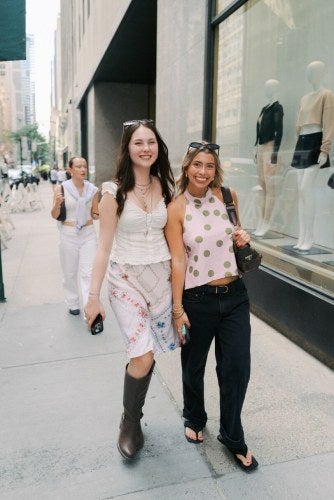 two young female students walk down 5th Avenue in New York City by a fashion business window display.