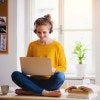 A woman sitting on a tabletop has a laptop on her lap. She is wearing headphones and a yellow shirt. 