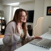 online student in glasses and sweater sitting at laptop at home, waving 